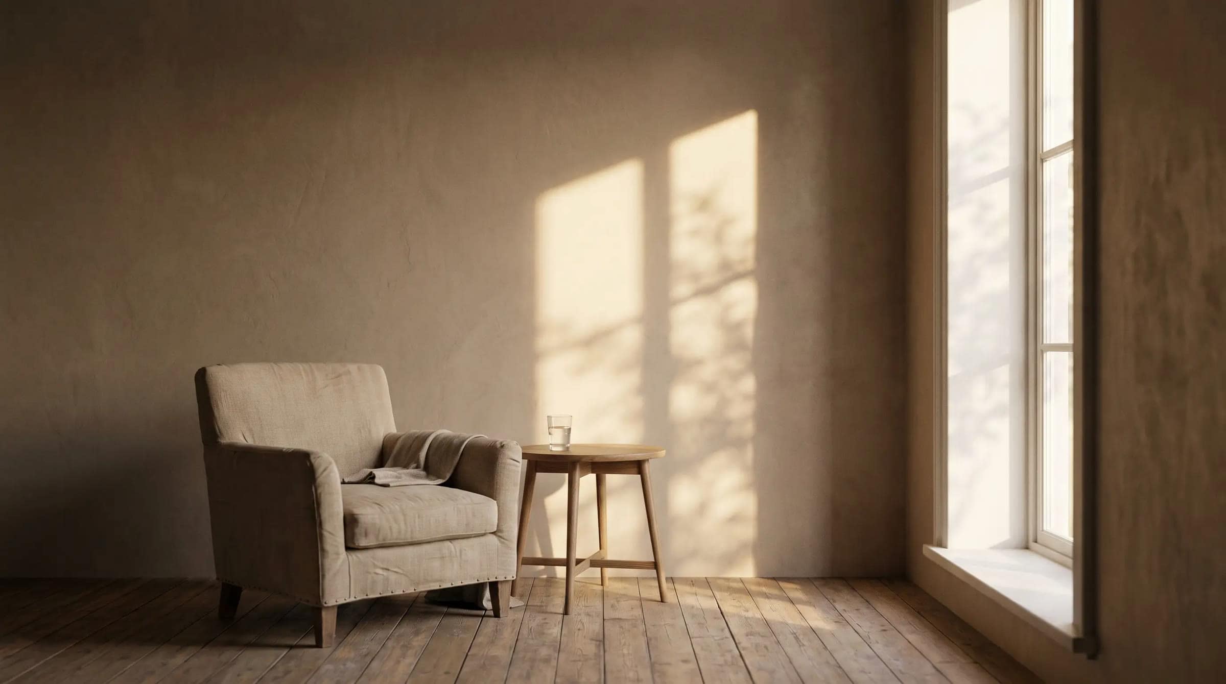 A quiet room in the morning: a linen armchair and a small table with a glass of water, and sunlight with the shadow of leaves on a warm plaster wall.