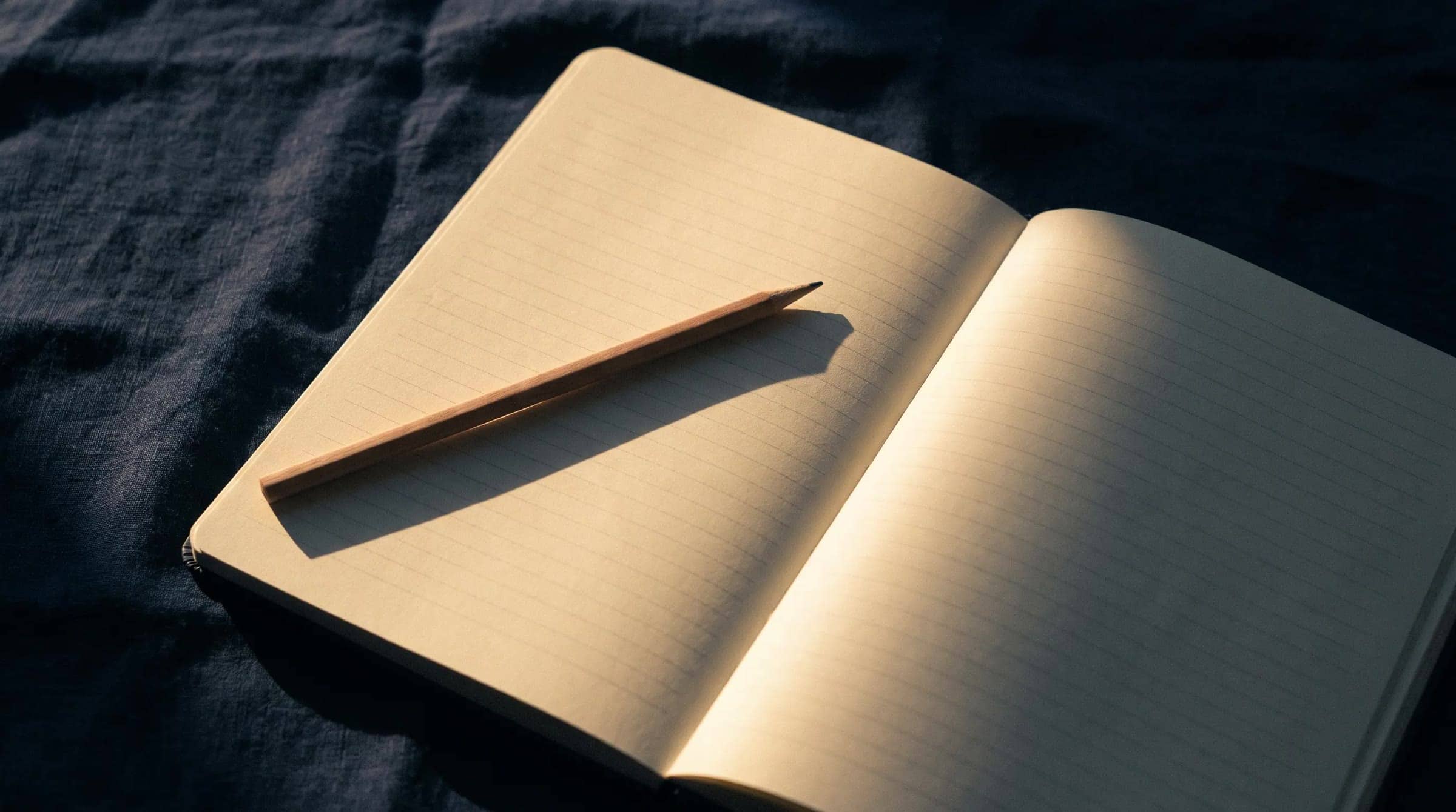 A blank workbook lying open on indigo linen, a pencil across its page, in low morning light.
